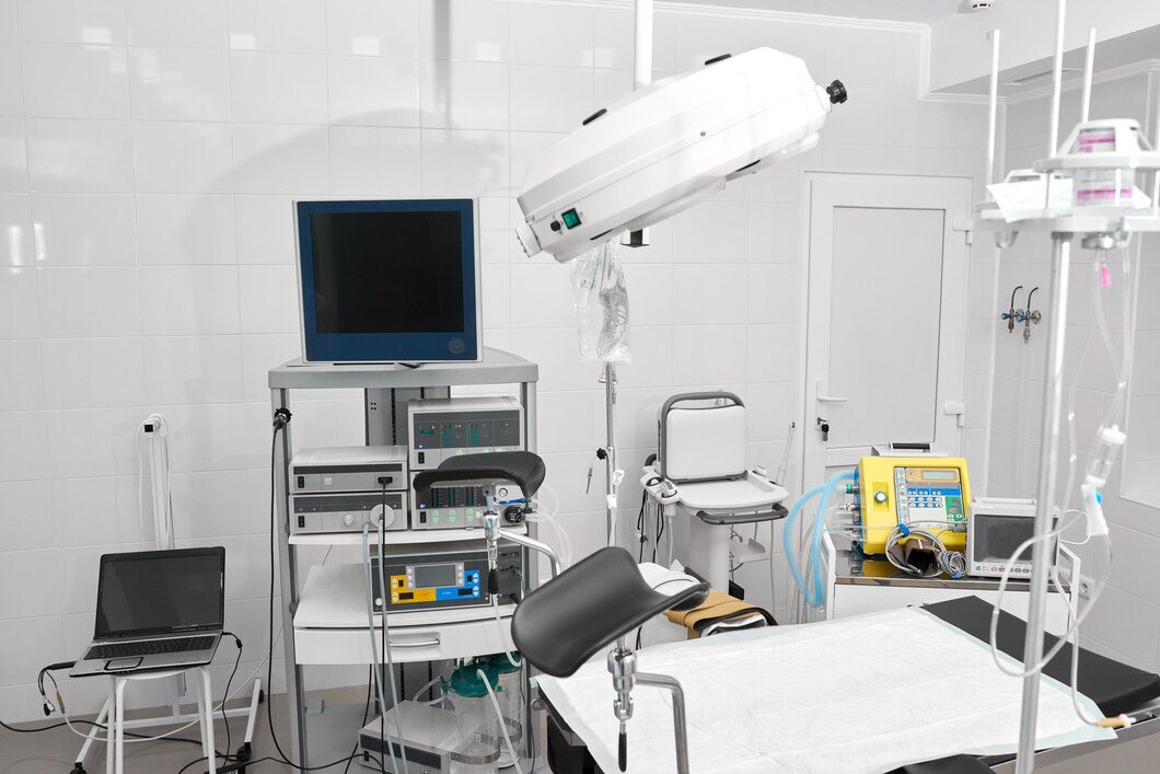 A modern hospital operating room equipped with various medical devices, including monitors, a ventilator, surgical lights, and other electronic equipment. An adjustable examination table with a white sheet is positioned in the center.